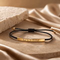 Black cord bracelet with gold bar and 'God Is In Control' engraving on a beige fabric background